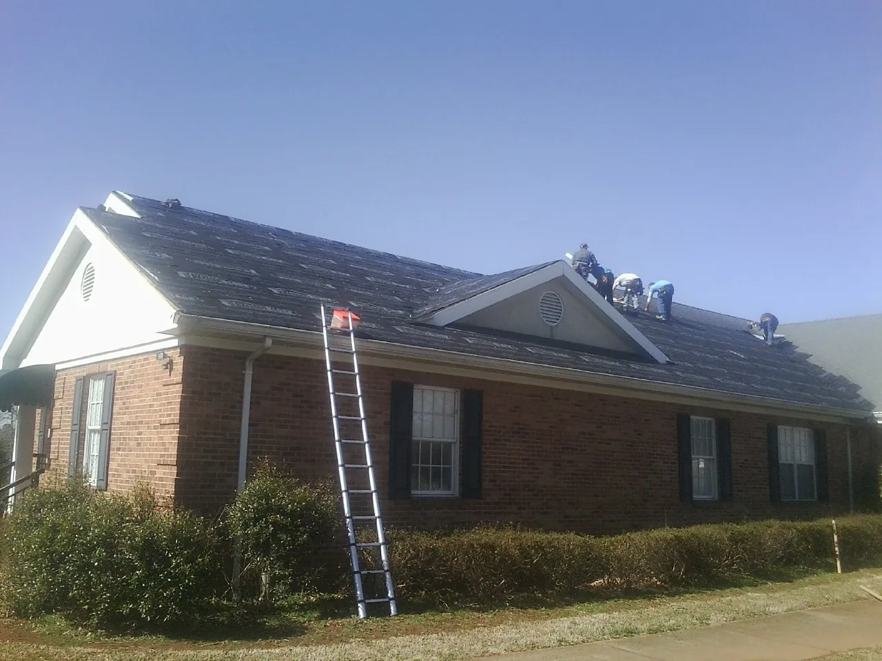 Crew tearing shingles off a brick ranch roof with a ladder set — Emergency Roof Repair work by our crew in Sunset Beach