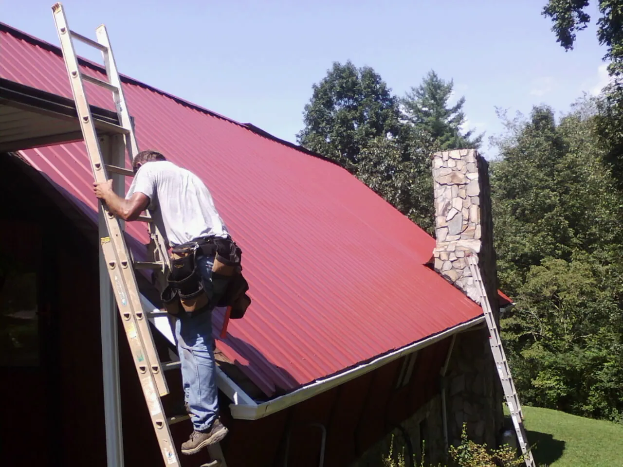 Roofer on a ladder beside a red metal roof and stone chimney — Soffit & Fascia Repair by Roofing Brothers in Sunset Beach